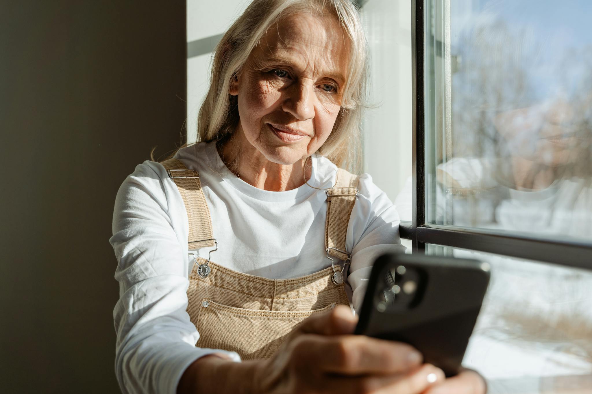 A senior woman enjoys leisure time browsing on her smartphone by a sunlit window.